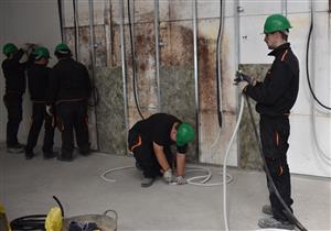 Five men in working uniforms are wearing green helmets. One of them in kneeling on the floor working with a cable. A man to his right holds together two tubes.
