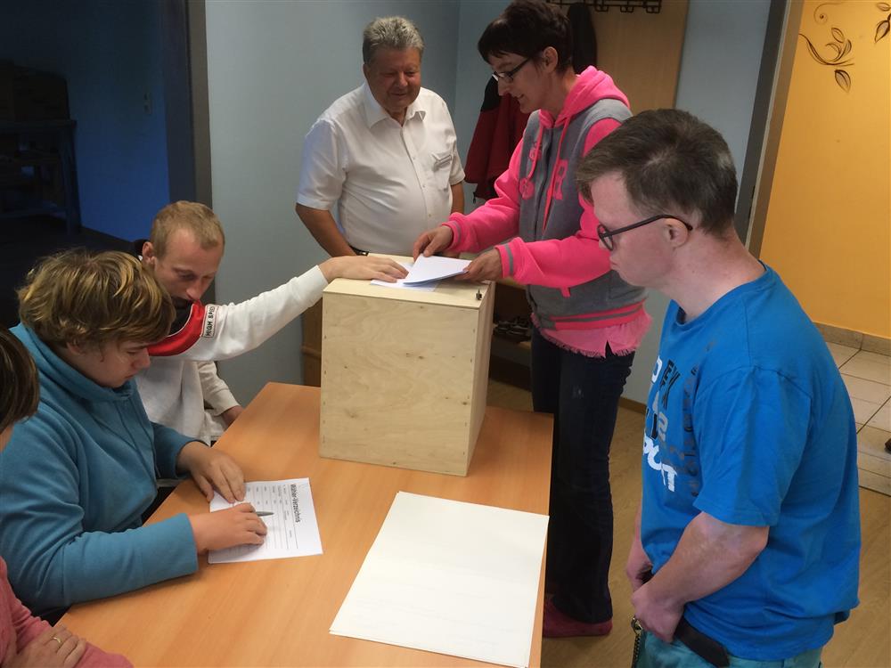 A man who appears to have down syndrome standing in front of a woman checking a list. One man looking at the list with his hand is on top of the box while two other individuals are discussing beside him.