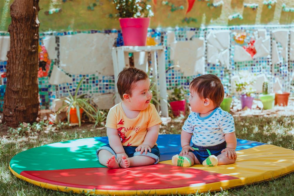 The image captures a tender moment between two young children sitting on a colorful play mat outdoors. The mat features vibrant sections of red, green, blue, and yellow, and the children are engaging with each other, suggesting a sense of curiosity and friendship. The background is a garden setting with a variety of plants and decorative elements, creating a peaceful and playful environment. The children's expressions and body language reflect innocence and the universal nature of childhood, transcending any specific cultural or geographic origin. The scene promotes themes of equality and tolerance, as the children interact without prejudice, exemplifying the pure and accepting nature of early childhood interactions.