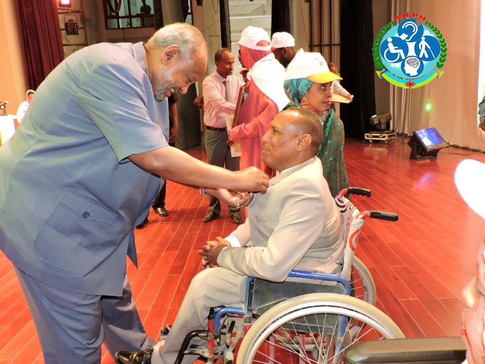 A man puts on a pin on the shirt of an elderly man in wheelchair while people lining up behind them.