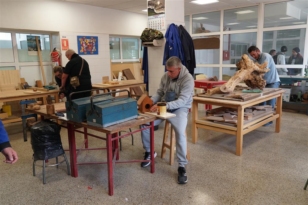 There are several men working on wood. One of them has built a birdhouse, and one man in the background works on a tree trunk. The setting is in a carpenter shop with tools and work benches.