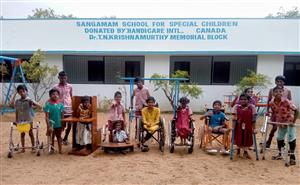 A group of Indian children in different ages appearing to have different motor disabilities assisted by crutches and wheelchairs are in the playground in front of the Sangamam School for Special Children building.