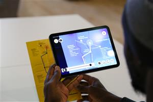 A person uses a tablet to view an interactive digital map, overlaying information onto a printed guide. The scene illustrates how accessible technology bridges information and navigation, empowering users with visual or cognitive support.