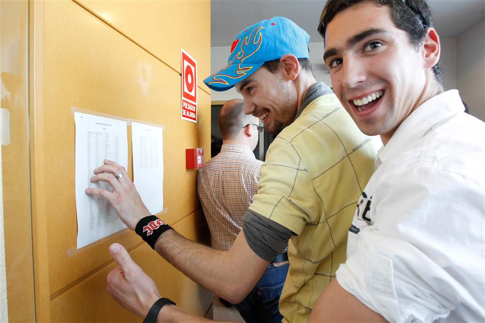 Two young men, one in a white T-shirt and one in a yellow T-shirt wearing a blue hat look at a piece of paper on the wall listing the exam rooms. Both are smiling and laughing.