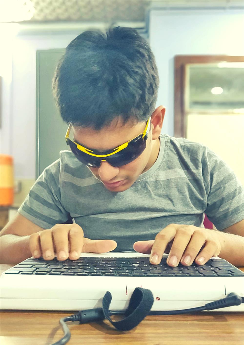 A young person with dark glasses is typing on a keyboard in front of him. He seems to be very concentrated.