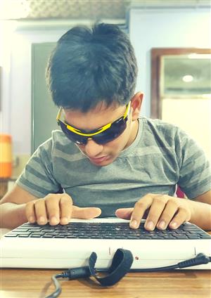 A young person with dark glasses is typing on a keyboard in front of him. He seems to be very concentrated.