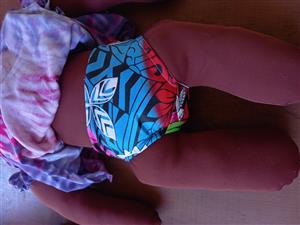 A handmade doll with dark brown fabric skin, colorful patterned shorts, and a tie-dye cloth lies on a table. The doll’s design celebrates cultural identity and representation, emphasizing the importance of inclusion and diversity in children’s toys.