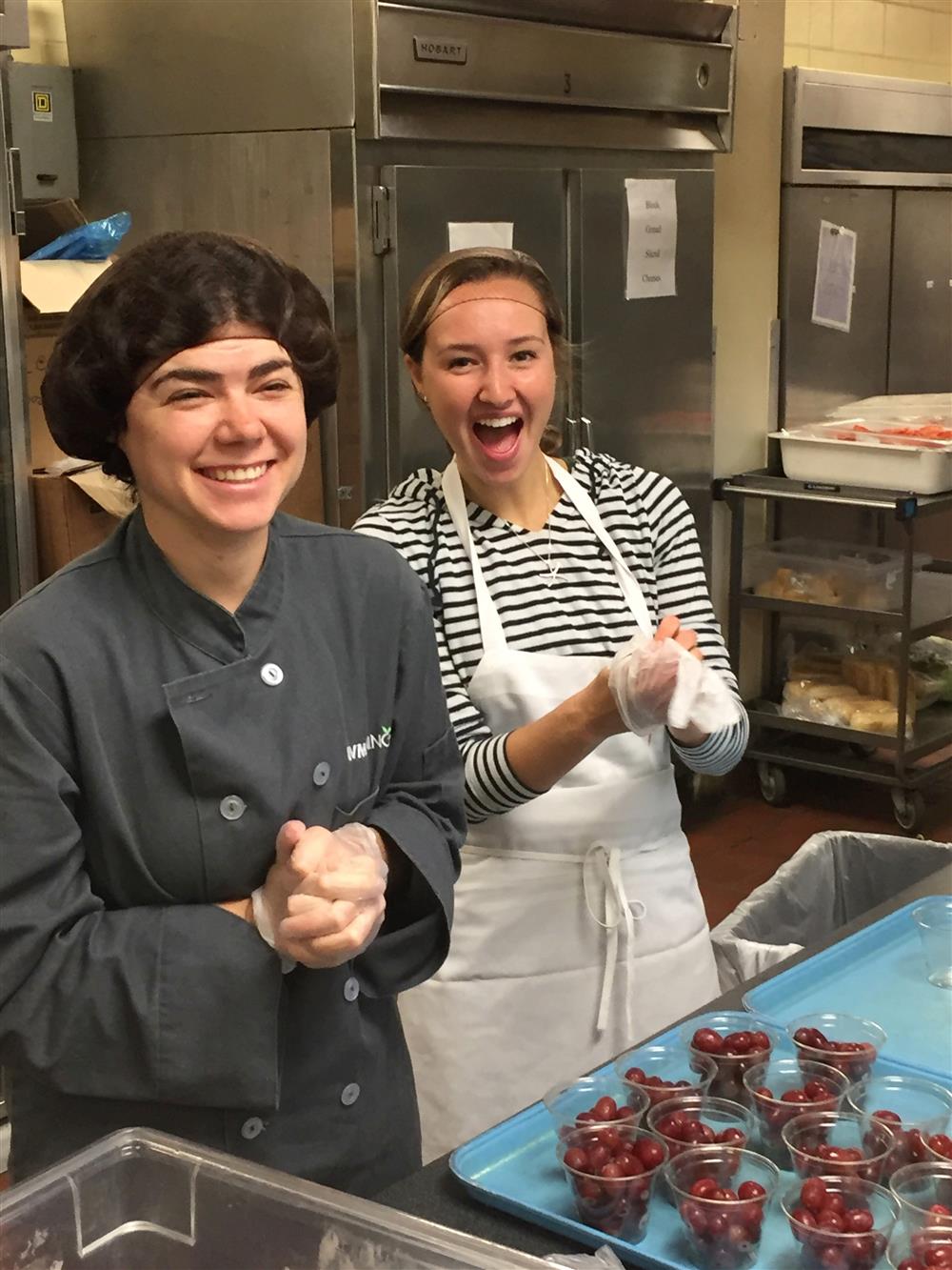 Two women stand in a catering kitchen, laughing and smiling. Full cups of grapes are arranged on a tray in front of them.