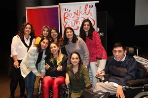 The image shows a joyful group of nine individuals posing together for a photo. Two individuals are using wheelchairs, which signifies the inclusion of people with disabilities. The group is diverse in terms of gender and possibly cultural backgrounds, reflecting a spirit of equality and tolerance. Behind them, a banner with vibrant colors and the words "Renkli Kampus" suggests a context of community, possibly related to an educational or social initiative that promotes diversity and inclusion. The smiles and positive body language of the individuals convey a message of camaraderie and support for one another.
