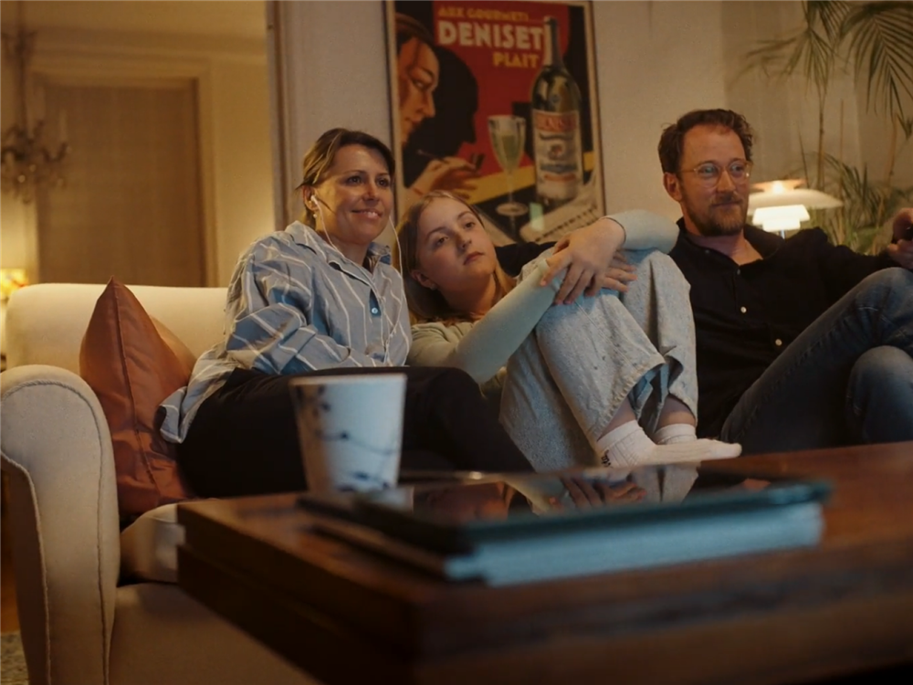A family of three, one daughter, father and mother are sitting in front of the television with the mother receiving the audio-translation of the subtitles via earphones connected to the Subreader app.