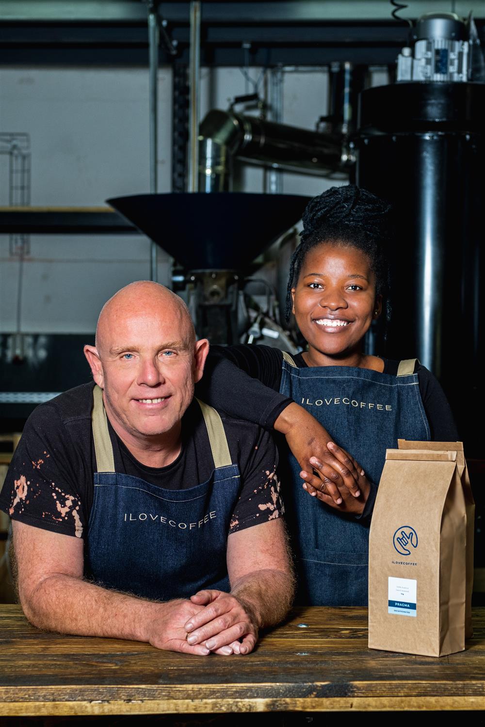 A young girl and a man in I Love Coffee uniform lean against a counter; two coffee bags are next to them. They are smiling 