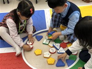 The photo shows three young children engaged in a playful activity on the floor. They are surrounded by colorful educational toys, such as puzzle pieces and blocks with numbers and letters on them, suggesting a learning environment. The children appear to be Asian and are dressed in casual, comfortable clothing. One child is focused on placing a puzzle piece, another is holding a toy, and the third is reaching out to a piece, indicating cooperation and shared playtime. The setting promotes themes of learning, equality, and the importance of nurturing environments for children's development.