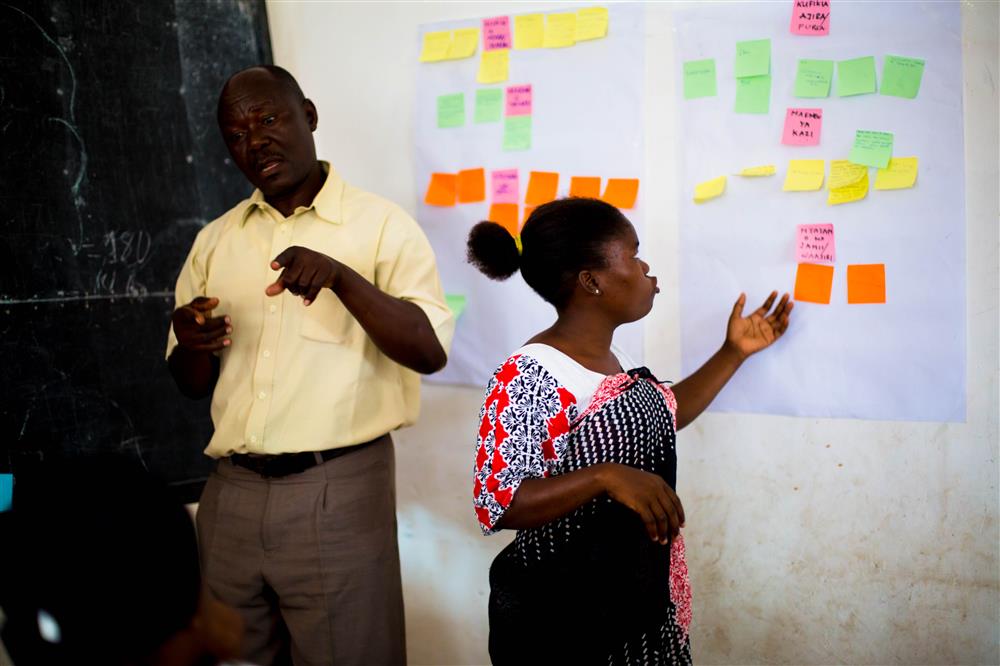 A woman stands in a classroom and points to a sheet of paper on the wall with lots of colourful sticky notes on it. A male sign language interpreter stands to the left of her facing the room.