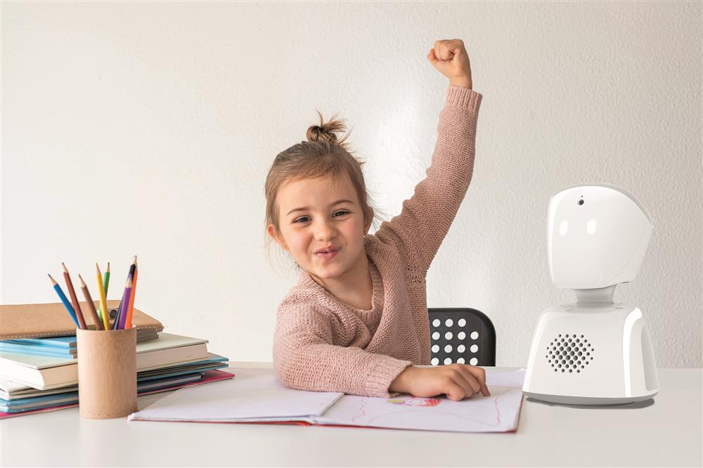 A preschool girl seated on her desk with left arm raised. On her left is a robot and on her right are books piles and a canister with pencils. 