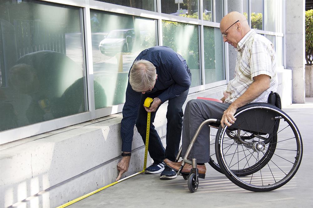 One old men and a men in a wheel chair measure the floor outside a building, in order to improve its accessibilty and becoming trained RHFAC Professionals.