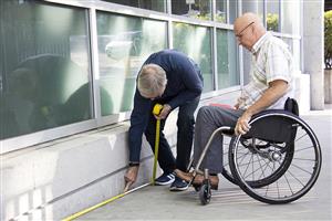 One old men and a men in a wheel chair measure the floor outside a building, in order to improve its accessibilty and becoming trained RHFAC Professionals.