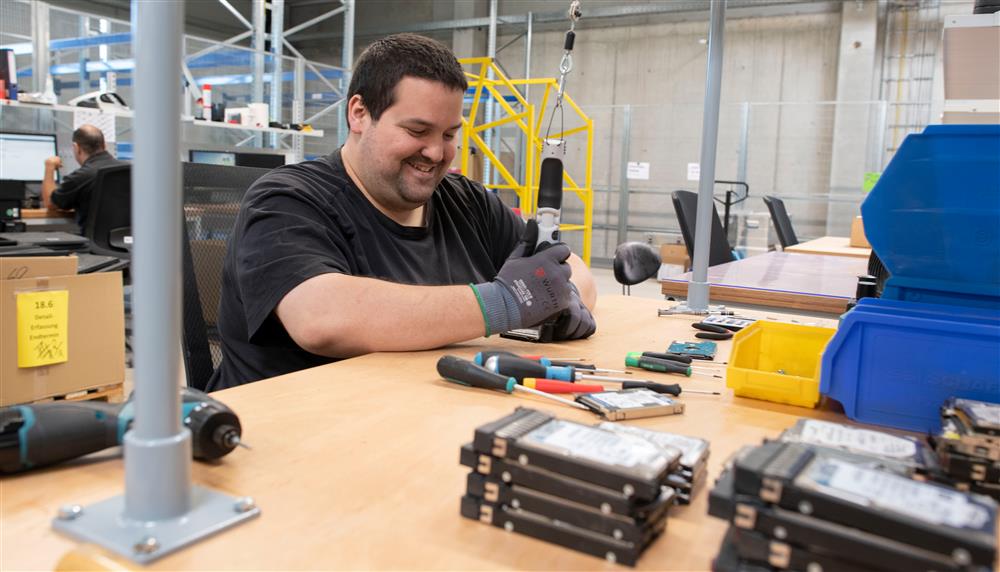 A smiling man sits at a workspace in an industrial workplace. The man is holding a tool and is working on a hard drive on the desk in front of him. 
