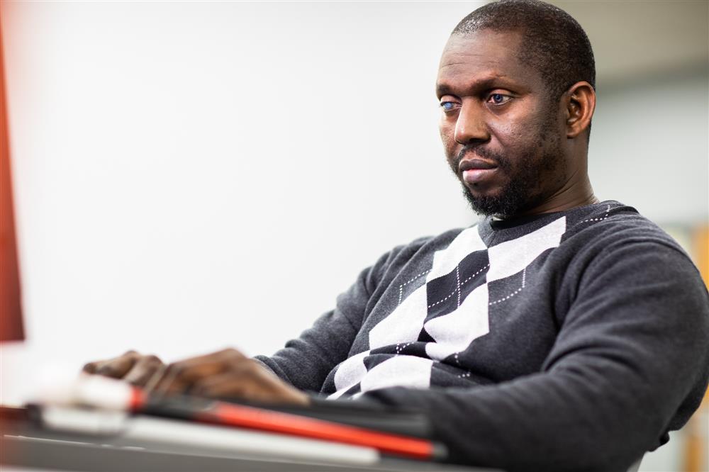 The photo shows an individual who appears to be a man with a focused or contemplative expression. He is wearing a sweater with a geometric pattern and seems to be seated at a desk or table, suggesting a work or study environment. The background is out of focus, emphasizing the person in the foreground. There are no explicit elements in the image that denote themes of equality, tolerance, assistance, or justice, but the setting might imply a commitment to education or professional development, which can be associated with personal empowerment and opportunity.
