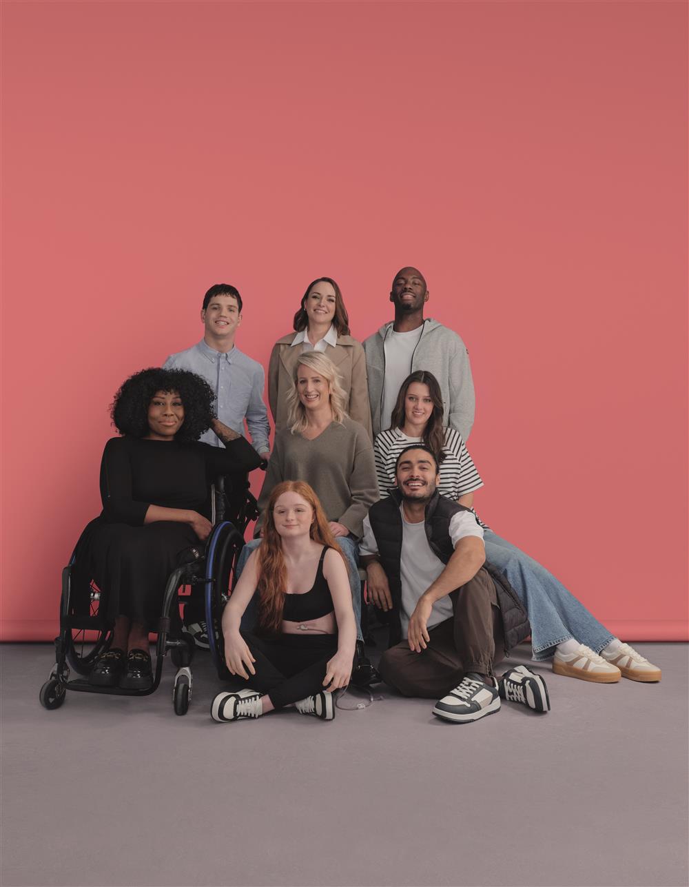 A group portrait of eight diverse individuals against a pink background. Among them is a woman using a wheelchair, and the group includes people of various genders and appearances. Their relaxed, happy expressions convey a message of belonging, empowerment, and inclusive representation.