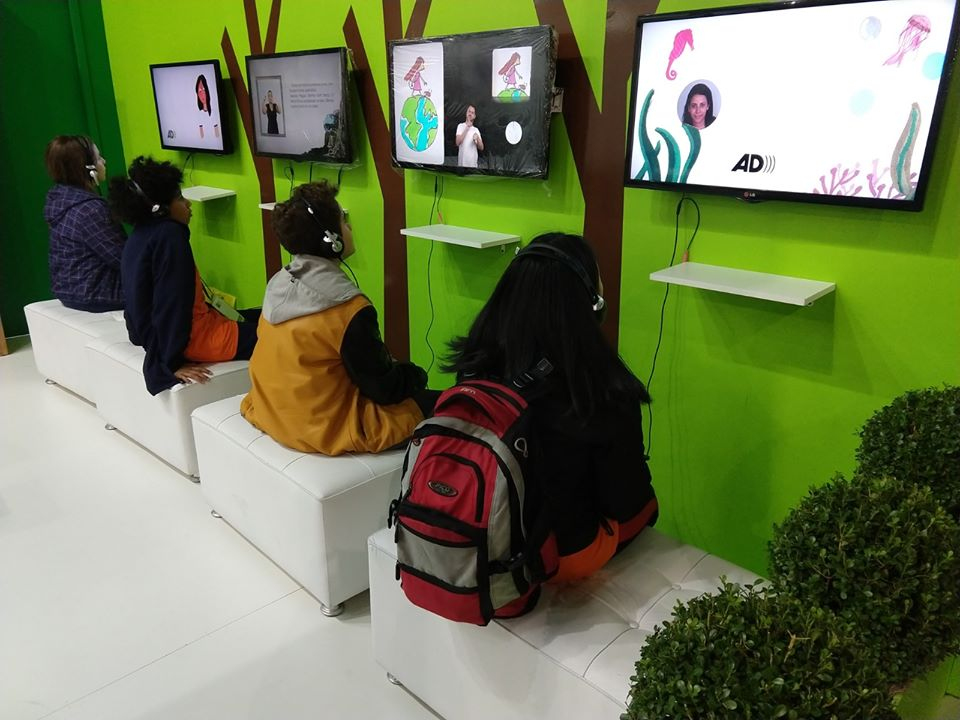 Four women sitting on white couches in front of screens showing persons and graphics. Each of them has headphones on. The screens hang on a bright green wall with trees drawn upon them. 