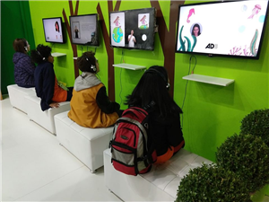 Four women sitting on white couches in front of screens showing persons and graphics. Each of them has headphones on. The screens hang on a bright green wall with trees drawn upon them. 