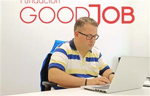 In the image, we see an individual focused on working at a laptop. The person is wearing glasses and a striped polo shirt, and appears to be sitting in an office environment. Behind them is a wall with the words "Fundacion GOODJOB" prominently displayed, suggesting that this may be a workspace of a foundation that likely focuses on social good, employment, or assistance. The setting conveys a professional atmosphere, and the person's engagement with their work could be seen as a representation of inclusion and empowerment in the workplace.