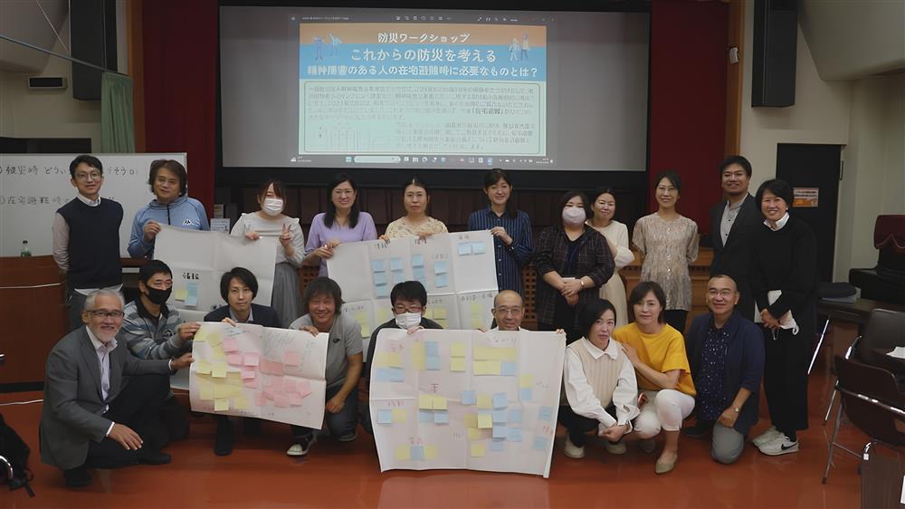 A diverse group of people in Japan poses after a workshop on disaster preparedness for individuals with disabilities. They hold colorful posters filled with notes, symbolizing collaboration and inclusive planning that ensures everyone’s safety and dignity in emergencies.
