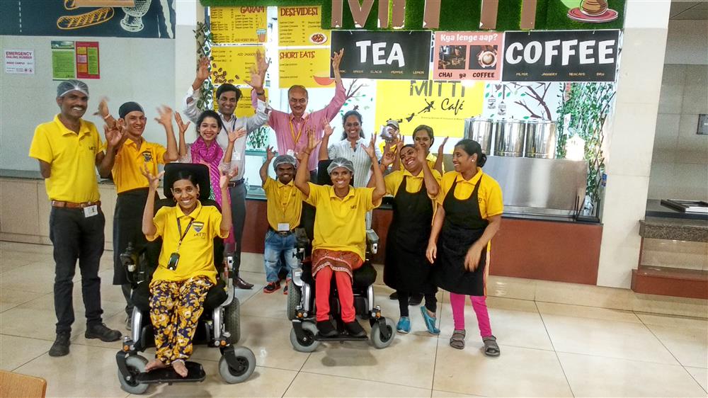 A group of 11 people in front of a café serving counter. Two people at the front of the group are in wheelchairs, the rest are standing and all are smiling towards the camera and waving.