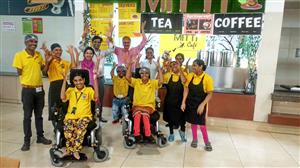A group of 11 people in front of a café serving counter. Two people at the front of the group are in wheelchairs, the rest are standing and all are smiling towards the camera and waving.