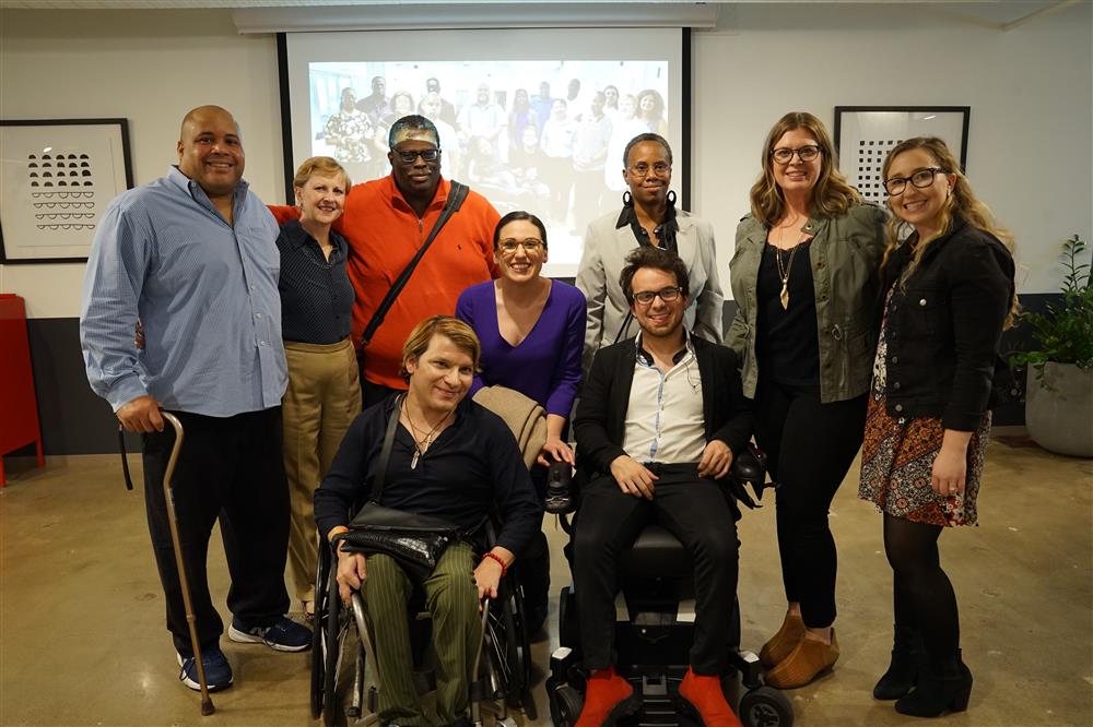 A group of men and women of diverse backgrounds and abilities gather together, some using wheelchairs or other mobility aids. Their expressions are warm and collaborative, embodying unity, respect, and inclusivity. This scene highlights a supportive environment where diverse abilities and experiences are valued.