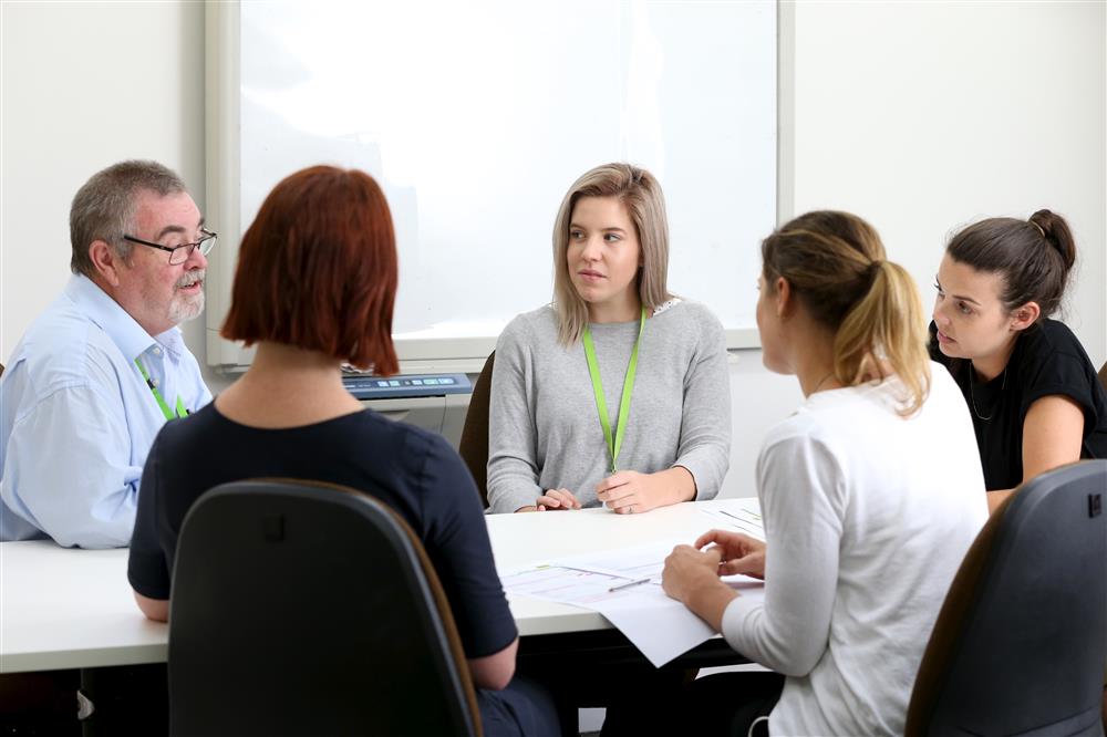 Four women and one man sit at a table in a meeting room. Two of them have their backs to the camera. The man (left of the picture) is talking and the other people at the table are looking at him.