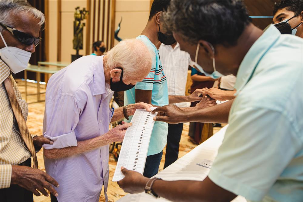 A Sri Lankan man appearing to be blind holding the elbow of an elderly man who is looking at a lengthwise paper printed with different shapes held by another man. A group of men discussing behind them. 