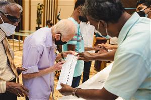 A Sri Lankan man appearing to be blind holding the elbow of an elderly man who is looking at a lengthwise paper printed with different shapes held by another man. A group of men discussing behind them. 