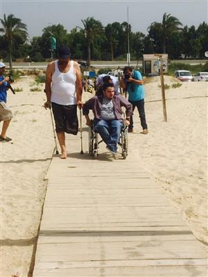 Two men with physical disabilities walking on a wooden platform on a sandy ground to reach the sea.