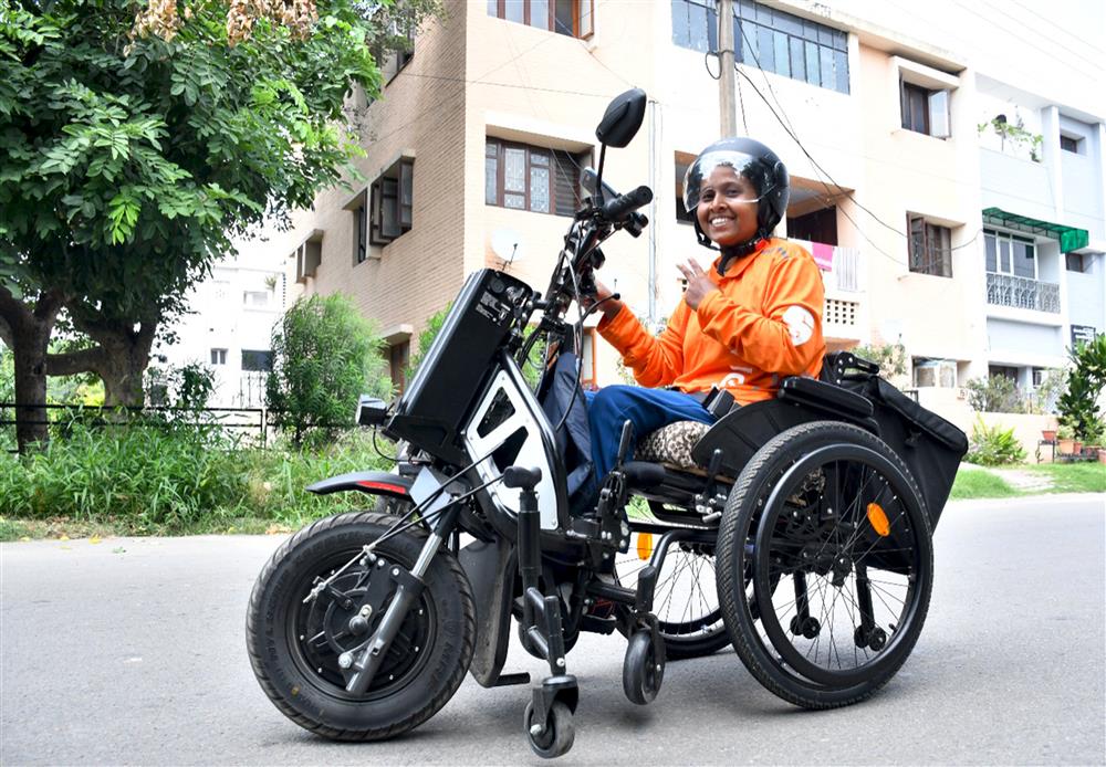 A woman, likely from South Asia, is shown on a wheelchair-accessible motorcycle, conveying freedom and empowerment. Her bright orange shirt and helmet suggest safety and preparedness. This image promotes mobility and independence for people with disabilities, highlighting adaptive technology's impact on inclusivity.