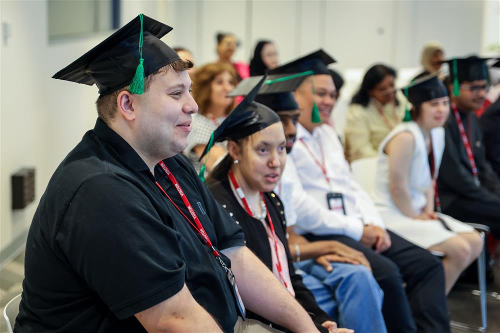 The photo shows a diverse group of individuals seated in what appears to be a graduation ceremony, as suggested by the academic caps they are wearing. The focus is on two people in the foreground who seem to be experiencing a moment of joy and pride. The individuals come from various backgrounds, reflecting a multicultural environment. Their expressions convey a sense of accomplishment and happiness. The setting promotes themes of achievement and inclusivity, as it captures a moment where personal efforts are celebrated, regardless of one's background or abilities. The atmosphere is one of positivity and mutual support, aligning with values of equality, tolerance, and justice.