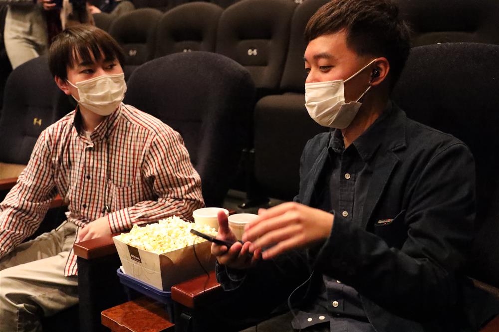 Two men both wearing surgical mask seated inside a cinema with a bucket of popcorn and two cups of drinks in between them. One of the men is holding a mobile phone and headphones in his ears.