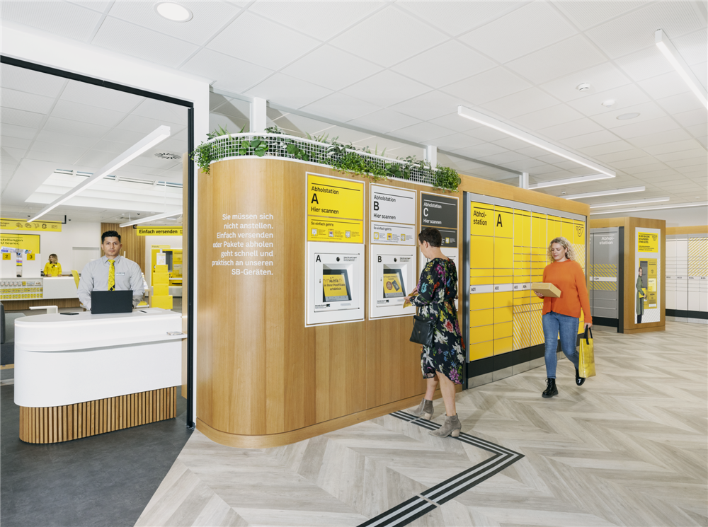 An accessible mail pickup station is shown in this photograph. Two women are picking up parcels, one walking and one standing in front of a touch screen, while a service employee awaits them on the left.