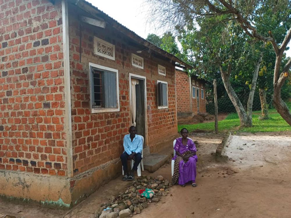 An African man and African woman sitting outside a well-built brick house with two windows and a door. A stack of rocks scattered between the with a tote bag lying on top. 