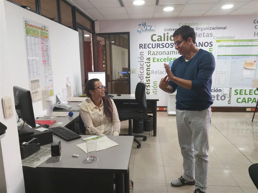 A young woman with dark hair sits at a desk in an office. She is talking to a man standing next to her.