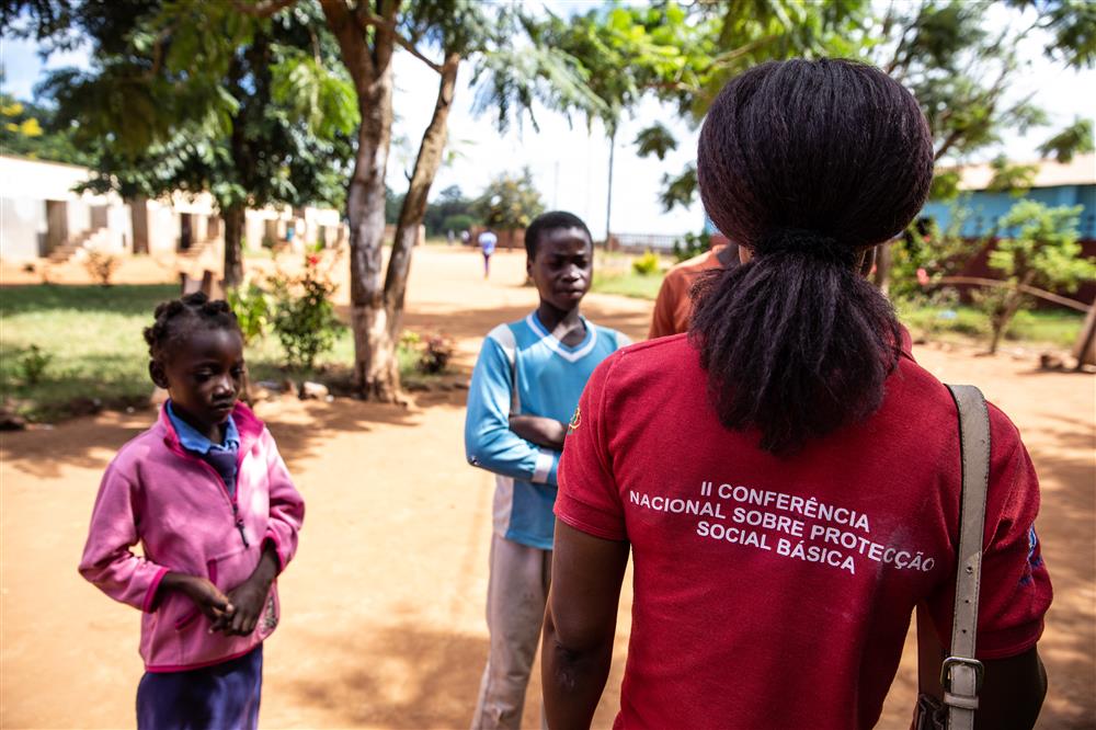 A teacher wearing their company tshirt is facing two children, who are getting inclusive education outside in the shadow.