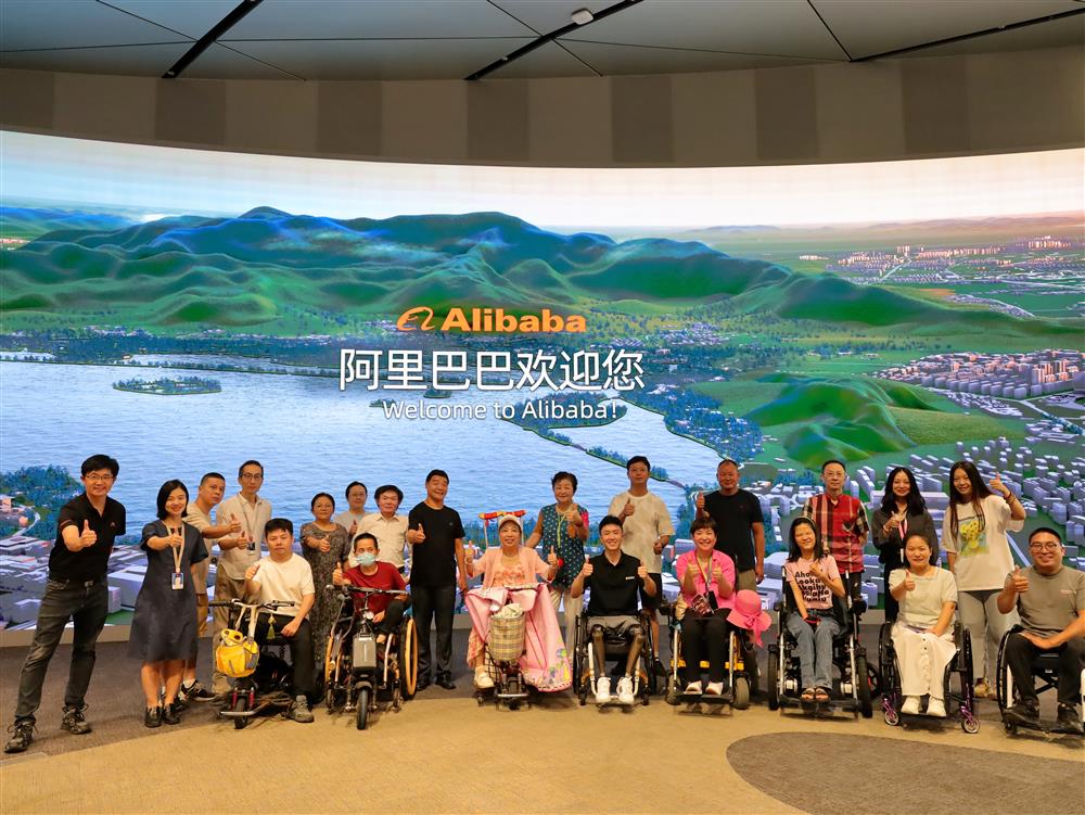 A diverse group of people, including wheelchair users, stand together smiling in front of a large digital screen at Alibaba headquarters. The scene represents inclusion, accessibility, and empowerment in the workplace, celebrating equal participation in technology and innovation.