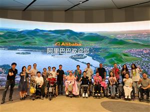 A diverse group of people, including wheelchair users, stand together smiling in front of a large digital screen at Alibaba headquarters. The scene represents inclusion, accessibility, and empowerment in the workplace, celebrating equal participation in technology and innovation.