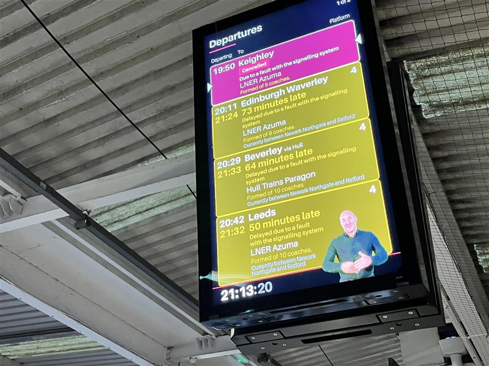 A train station departure board displays travel updates with a sign language interpreter shown on screen. This thoughtful inclusion of accessibility features ensures equal access to information for Deaf travelers, representing progress toward inclusive public spaces.