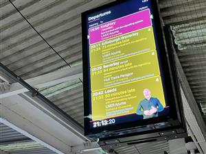 A train station departure board displays travel updates with a sign language interpreter shown on screen. This thoughtful inclusion of accessibility features ensures equal access to information for Deaf travelers, representing progress toward inclusive public spaces.