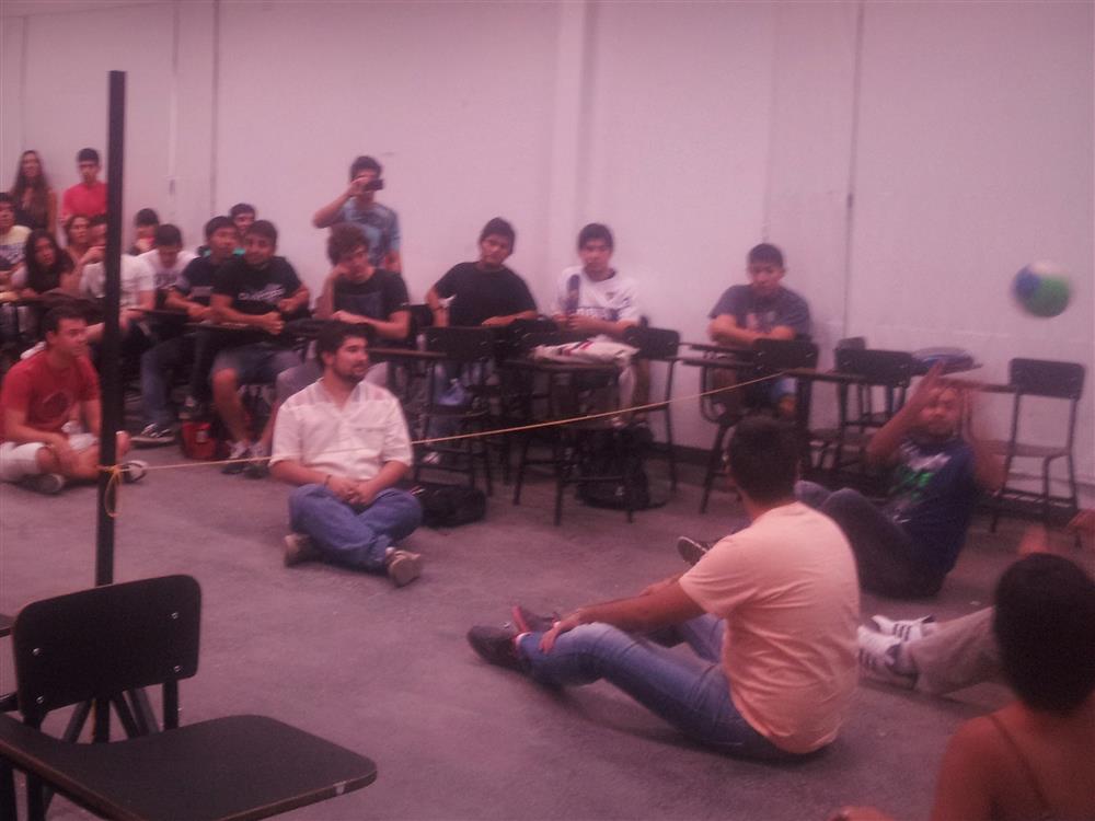 A classroom setting in a universtity. A group of young male students sit on the floor in an imporvised playing field trying to throw a ball over a slim cord, dividing the field. Their classmates cheer and take fotos in the background..