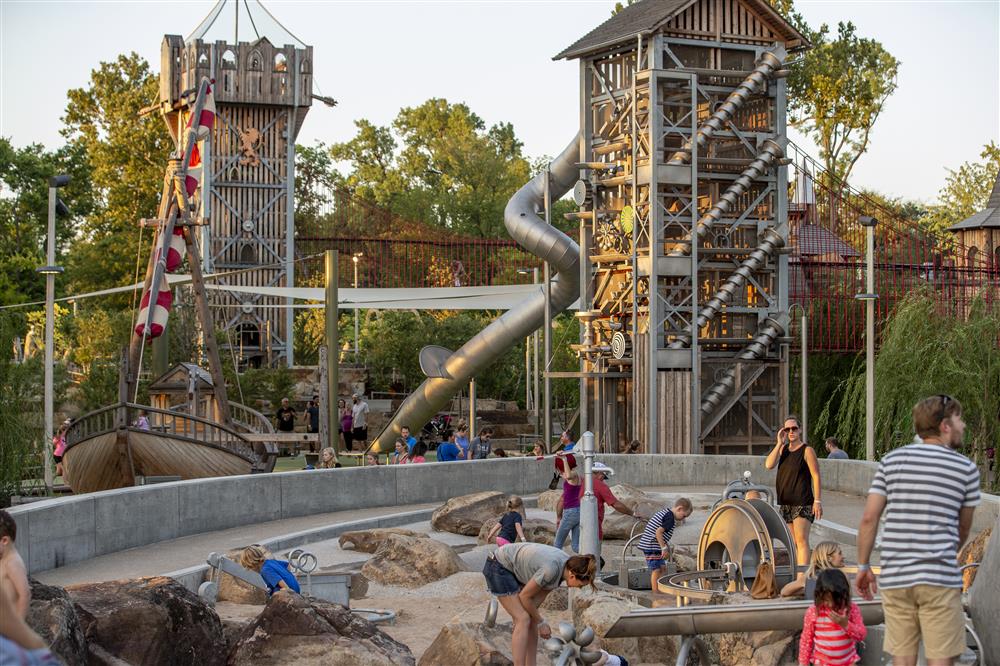 This image depicts an enormous accessible entertainment park with two huge towers and a silde, a long red bridge, a pirate boat and children playing in a sandpit with their parents. 
