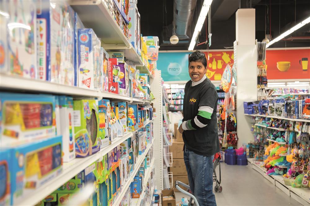 A  woman is standing on a step ladder in front of a shelf filled with children's toys inside a supermarket while smiling.