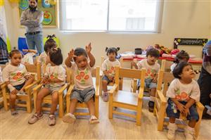 This image features a group of young children sitting in wooden chairs, most likely in a classroom or daycare setting. They are wearing matching white t-shirts with a colorful design, and their expressions range from curious to joyful. Some children are looking towards the camera, while others are engaged with their surroundings. In the background stands a man, arms crossed, observing the children—possibly a teacher or caretaker. The room has a cheerful ambiance with decorations on the walls, and there's a table with various items, suggesting an environment for play and learning. The scene embodies themes of community, nurturing, and the innocence of childhood.