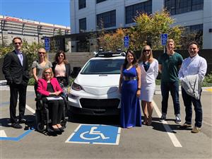 3 men and 5 women, one of whom is sitting in a wheelchair, are positioned around a modern white car with sensors on the roof, standing on a parking space for persons with disabilities. 